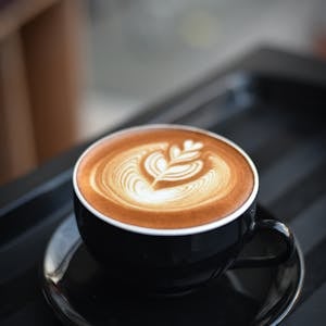 A beautifully crafted cappuccino with intricate latte art in a black ceramic mug on a dark surface.